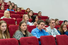 A group of people sitting in the auditorium in rows on red chairs. The people in the photo are of different genders and ages. Most of the participants are focusing their attention on the event taking place outside the frame - some of them have open notebooks or notepads. A plain white wall is visible in the background.