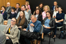 A group of people sitting on chairs in a conference room. The participants are dressed formally - in suits, jackets, elegant blouses and shirts. Some people are holding notepads, pens or cups of coffee in their hands. Most of them are facing the meeting leader, who is out of the frame of the photo. The background is a wooden wall and a section of a darker wall.