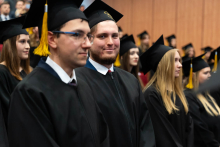 Grupa osób uczestniczących w ceremonii. Uczestnicy stoją w kilku rzędach i są ubrani w tradycyjne czarne togi oraz birety z żółtymi chwostami. W tle widoczni są inni uczestnicy wydarzenia, również siedzący, a całość odbywa się w przestronnej auli z drewnianą ścianą w tle.