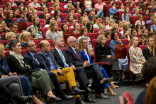 Duża grupa osób siedzi w audytorium na czerwonych krzesłach. W pierwszym rzędzie widocznych jest sześć osób, w tym mężczyzna w żółtych spodniach i kobieta w niebieskim garniturze.