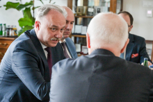 Czterech mężczyzn siedzących przy stole. Dwóch mężczyzn jest widocznych od tyłu, dwóch od przodu. Mężczyzna po lewej stronie ma siwe włosy i nosi granatowy garnitur. Mężczyzna po prawej stronie ma siwe włosy i nosi ciemny garnitur. W tle znajduje się zielona roślina doniczkowa. Obok rośliny stoi drewniana komoda z różnymi przedmiotami na blacie.