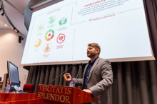 Mężczyzna stojący przy mównicy, wygłasza prezentację w audytorium. Na mównicy widnieje napis 'VERITATIS SPLENDOR'. Za mężczyzną znajduje się duży ekran projekcyjny, na którym wyświetlane są slajdy z tekstem i grafikami.