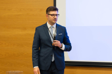 The middle-aged man is dressed in a smart navy blue suit, white shirt and checkered tie. The man is standing against a wooden wall and a projection screen. He holds a microphone in his right hand, while his left arm falls freely along his body. Around his neck hangs an ID badge. 