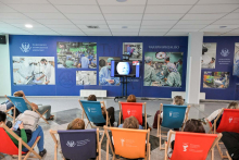 Grupa osób siedzi na kolorowych leżakach w sali konferencyjnej, oglądając prezentację na trzech ekranach. W tle zdjęcia z sal operacyjnych i logo Warszawskiego Uniwersytetu Medycznego. Ściany ozdobione plakatami uczelni.