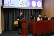 Po lewej stronie przy brązowej mównicy stoi kobieta. Jest ubrana w ciemną sukienkę, ma okulary i ciemne włosy. Za mównicą znajduje się ekran projekcyjny. Po prawej stronie siedzą dwie kobiety przy pulpicie konferencyjnym, na którym znajdują się komputer i butelki z wodą. W tle, na ekranie projekcyjnym, widoczne są cztery logotypy różnych organizacji lub instytucji.