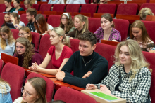 A group of young people sitting in rows on red chairs. Three people are visible in the foreground: a woman in a red short-sleeved blouse, a man in a black sweater, and a woman in a black-and-white patterned jacket. Further down the room, more participants can be seen sitting and looking ahead or reviewing their notes.