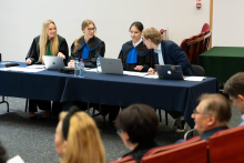 Cztery osoby siedzą za stołem przykrytym granatowym obrusem. Są to trzy młode kobiety i jeden młody mężczyzna. Kobiety mają na sobie togi. Mężczyzna w marynarce pochyla się w stronę osoby obok, jakby coś jej mówiła. Na stole znajdują się trzy laptopy, kilka butelek wody oraz dokumenty.