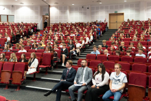 Sala wypełniona ludźmi siedzącymi na czerwonych krzesłach. Na pierwszym planie, w dolnej części zdjęcia, znajduje się kilka osób siedzących na krzesłach, ubranych formalnie i nieformalnie.