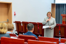 A middle-aged woman, dressed in a light gray suit, standing in a conference room and speaking to an audience. The woman holds a microphone in her right hand and a piece of paper in her left. In front of her, in several rows, listeners sit on red chairs with wooden backs - the backs of their heads are mostly visible. In the background, to the right, is a lectern and draped gray curtains.