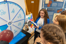 Grupa osób stojących wokół dużego koła fortuny z różnymi symbolami. Koło jest podzielone na sekcje, każda z nich zawiera inny symbol, takie jak stetoskop, mikroskop, kolba laboratoryjna i inne. Obok koła stoi osoba trzymająca kartki papieru i rozmawiająca z grupą.