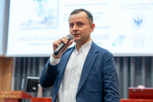 A middle-aged man wearing a white shirt and blue jacket. The man holds a microphone in his right hand and speaks into it. In the background you can see a projection screen with logos of various institutions. 