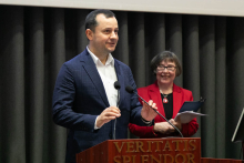 Po lewej stoi mężczyzna. Jest ubrany w granatową marynarkę i białą koszulę, ma ciemne włosy. Stoi za mównicą i przemawia. Po prawej stoi kobieta, która ma na sobie czerwoną marynarkę. W tle widoczna jest ciemna zasłona.