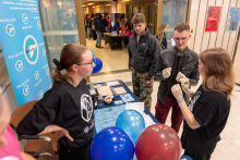 Grupa młodych ludzi stojących przy stoliku na Nocy Muzeów. Na stoliku znajdują się różne materiały, takie jak ulotki, długopisy i balony w kolorach niebieskim i czerwonym. W tle widoczne są inne stoiska oraz osoby uczestniczące w wydarzeniu.