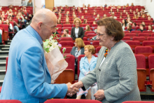 The auditorium during the conference. In the foreground, on the left, stands a man dressed in a light blue jacket, holding a bouquet of flowers wrapped in paper. The man shakes hands with the woman standing across from him. The woman is dressed in a gray jacket and has a colorful necklace around her neck. In the background can be seen rows of red upholstered chairs on which the participants of the event are seated. Most of the seats are occupied, and people are looking toward the stage.