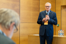 An older man in a suit standing in front of a light wooden wall and speaking into a microphone. The man holds the microphone in one hand and has a name tag hanging around his neck. 