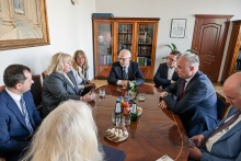 Grupa osób siedzących przy owalnym stole. Uczestnicy spotkania mają na sobie ciemne ubrania. Rozmawiają. Stół ma kolor brązowy. W tle widać regały z książkami i obraz na białej ścianie. 