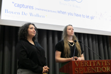 Dwie młode kobiety stojące za brązową mównicą. Kobieta po lewej stronie ma kręcone włosy i czarną bluzkę, a kobieta po prawej stronie ma długie proste włosy, czarną bluzkę i naszyjnik. Za nimi znajduje się ekran projekcyjny z tekstem.