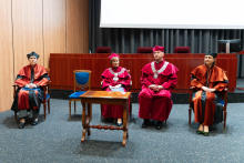 Grupa osób siedząca na fotelach na środku auli. Są to władze rektorskie i wydziałowe. Wszyscy są ubrani w togi i birety akademickie. Przed nimi stoi drewniany stolik. Za nimi widać fragment ekranu projekcyjnego oraz szare zasłony.