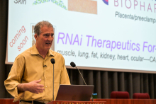 Mężczyzna w żółtej koszuli stoi za mównicą z mikrofonem i laptopem. W tle ekran z prezentacją na temat terapii RNAi.