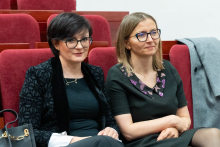 Dwie osoby siedzą w czerwonych fotelach w sali konferencyjnej. Osoba po lewej stronie ma krótkie ciemne włosy, okulary i nosi czarną marynarkę oraz czarną bluzkę. Osoba po prawej stronie ma jasne włosy do ramion i nosi czarną sukienkę z fioletowymi wzorami przy dekolcie.