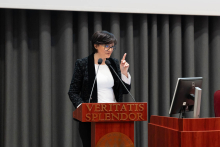 "Osoba stoi za drewnianą mównicą z napisem "Veritatis Splendor". Gestykuluje - ma uniesiony palec wskazujący. Jest ubrana w czarną marynarkę i białą bluzkę, mówi do dwóch mikrofonów przymocowanych do mównicy. W tle znajdują się ciemnoszare zasłony, a po prawej stronie mównicy stoi monitor komputerowy.