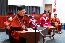 Na pierwszym planie znajduje się mężczyzna - dziekan Wydziału w pomarańczowo-czarnej todze, przemawiający do mikrofonu. Na nosie ma okulary. Na szyi ma ozdobny łańcuch i czarny biret na głowie. W tle, za mównicą, siedzi czterech innych uczestników uroczystości – również ubranych w togi i birety. Dwie osoby mają czerwone togi i białe łańcuchy, a pozostałe dwie – brązowe togi. 