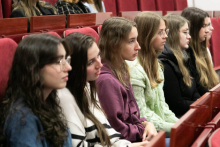 Grupa młodych kobiet siedzących w czerwonych fotelach audytoryjnych. Mają na sobie codzienne ubrania, takie jak swetry i kurtki. Na twarzach mają wyraz skupienia. Patrzą przed siebie.