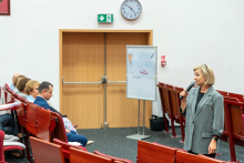 In the foreground, on the right, stands a middle-aged woman wearing a gray suit and holding a microphone. The woman is facing a group of people seated to the left of the photo, suggesting that she is speaking or giving a presentation.  In the background, behind the woman, is a board on a stand with the words “NURSES' DAY” and drawings depicting a nurse and a nurse's cap with a red cross. 