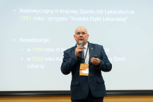 A middle-aged man wearing a navy blue jacket, light blue shirt and dark tie. The man stands in front of a projection screen, holding a microphone in his right hand and a remote control for a multimedia presentation in his left. Around his neck is a conference badge. The projection screen shows text in Polish on the history of the Code of Medical Ethics. In the background, on the left side of the frame, the blurred silhouette of another person is visible - an older man with gray hair.