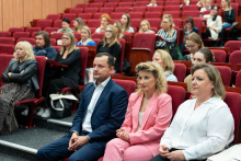 Grupa ludzi siedzących w audytorium na czerwonych krzesłach. W pierwszym rzędzie siedzą dwie kobiety i jeden mężczyzna. Mężczyzna siedzący po lewej stronie ma ciemny garnitur, kobieta siedząca w środku ma na sobie różowy garnitur, a kobieta po prawej stronie ma białą bluzkę. W tle widać więcej osób siedzących w rzędach za nimi.