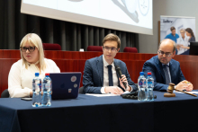 Trzy osoby siedzą przy stole przykrytym granatowym obrusem. Kobieta w białej bluzce i dwóch mężczyzn w granatowych marynarkach. Mężczyzna w środku jest młodszy i trzyma mikrofon w ręku. Na stole znajdują się butelki z wodą, laptop, mikrofon i młotek sędziowski. 