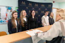 Grupę pięciu osób stoi za recepcją, są zwróceni twarzą do innej osoby po przeciwnej stronie blatu. W tle znajduje się ciemnoniebieski baner z logo i tekstem 'MIMESIS' powtarzającym się na całej powierzchni. Osoba po przeciwnej stronie blatu trzyma białą torbę z logo 'MIMESIS'.