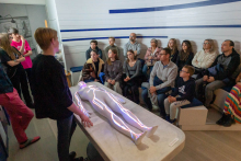 Grupa osób siedzących w półkolu, obserwujących prezentację. Na środku znajduje się stół, na którym leży szklany fantom człowieka. Fantom ma podświetlone linie przedstawiające układ krążenia. Dwie osoby stoją obok stołu i demonstrują na fantomie, jak zbudowany jest układ krążenia.