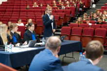 Sala  wykładowa z czerwonymi fotelami, w której odbywa się symulacja rozprawy sądowej. Przy stole znajdującym się po lewej stronie siedzą trzy kobiety w togach. Przy tym samym stole stoi mężczyzna i mówi do mikrofonu. Na pierwszym planie dwaj siedzący mężczyźni w garniturach widoczni od tyłu.