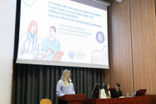 Po lewej kobieta osoba stojąca przy mównicy. Ma jasne włosy, okulary, jest ubrana w jasnoniebieską koszulkę. Obok za pulpitem konferencyjną siedzą dwie kobiety. Za nimi ekran projekcyjny, na którym wyświetlony jest slajd prezentacji z tytułem „Czynniki wpływające na decyzje kobiet o rezygnacji z kolejnej ciąży po pierwszym porodzie: analiza przyczyn i możliwości wsparcia”.