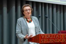 An elderly woman standing behind the lectern. She is dressed in a gray jacket, white blouse and a colorful necklace of large beads. The person's face has been blurred to protect privacy. Two microphones on long tripods are set up in front of the lectern. Gray curtains are visible in the background.