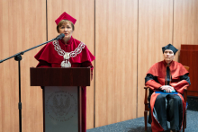 Po lewej stronie znajduje się kobieta - prorektorka, stojąca za drewnianą mównicą, przemawiająca do mikrofonu. Ubrana jest w czerwoną togę akademicką z ozdobnym łańcuchem rektorskim oraz czerwoną czapkę. W tle widoczna jest drewniana ściana. Po prawej stronie zdjęcia siedzi inna kobieta, ma na sobie togę. Za nimi jest drewniana ściana.