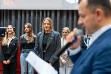 Four young women standing in a row on the auditorium stage. They are all facing the audience and looking ahead. In the background is a dark curtain and a section of a projection screen. In the foreground, to the right of the frame, is a man holding a microphone and a piece of paper. 