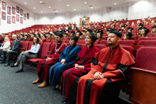Aula z czerwonymi fotelami, na której znajdują się uczestnicy dyplomatorium. Na pierwszym planie siedzą władze dziekańskie, ubrane w oficjalne togi w kolorach reprezentujących wydziały: bordowym, pomarańczowym oraz niebieskim.