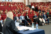 Sala wykładowa z czerwonymi fotelami, w której odbywa się symulacja rozprawy sądowej. Na pierwszym planie znajduje się stół przy którym siedzi kobieta w todze kilka  - jej twarz nie jest widoczna. W tle duża liczba uczestników siedzących na fotelach, patrzących w stronę mównicy.