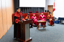 Grupa osób na auli. Na pierwszym planie stoi kobieta, prodziekan ds. farmacji, ubrana w pomarańczowo-czarną togę i czarny biret. Stoi za brązową mównicą i przemawia. Za nią na fotelach siedzi grupa osób ubranych w oficjalne togi akademickie. Togi są czerwone lub pomarańczowo-czarne. Za osobami widać ekran projekcyjny i szare zasłony.