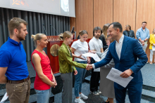 Presentation of certificates. On the stage of the auditorium, in two rows, stands a group of young people waiting to receive their certificates. A man in a blue suit is handing out the documents. A wooden lectern and a large projection screen are visible in the background. The room has wooden wall panels and a dark floor.