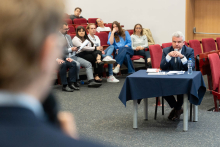 Na pierwszym planie, po lewej stronie, znajduje się osoba trzymająca mikrofon, której twarz jest rozmyta i niewidoczna. Na środku zdjęcia siedzi mężczyzna przy stole przykrytym granatowym obrusem, na którym znajdują się dokumenty i butelka wody. W tle widać grupę osób siedzących na krzesłach, niektóre z nich trzymają notatki lub urządzenia elektroniczne.