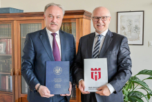 Dwóch rektorów stoi obok siebie. Mężczyzna po lewej trzyma niebieską teczkę z napisem „Warszawski Uniwersytet Medyczny” i godłem uczelni. Mężczyzna po prawej trzyma szarą teczkę z czerwonym logo. W tle znajduje się drewniana biblioteczka z książkami. Nad biblioteczką wisi obraz. Po prawej stronie zdjęcia stoi zielona roślina doniczkowa.