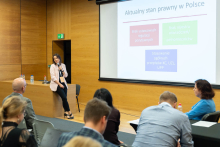 The woman is sitting on the edge of a desk in the conference room, holding a microphone and giving a presentation. Behind her is a screen with the title “The current state of the law in Poland” and three colored boxes with text. An audience sits in front of her, with some people taking notes.