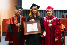 Trzy osoby pozujące do zdjęcia. Stoją na auli. Młoda kobieta w środku, absolwentka z najwyższą średnią, jest ubrana w czarną togę i biret. W rękach trzyma oprawiony dyplom. Po lewej stoi dziekan, po prawej prorektorka. Wszyscy się uśmiechają.