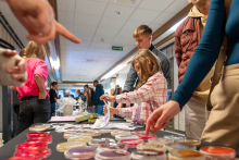 Stół z różnymi kolorowymi płytkami Petriego, które są ułożone na czarnym obrusie. Po prawej stronie stołu stoi dziewczynka w koszuli w kratę, która trzyma kartkę papieru i patrzy na płytki. Obok niej stoi dorosły mężczyzna w brązowej kurtce i beżowych spodniach. W tle widoczni są inni ludzie, niektórzy z nich rozmawiają lub oglądają wystawione przedmioty.