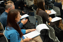 A group of people sits in a conference room. In the center, a woman in a blue shirt with a tattoo of two birds on her forearm holds a notebook and a pen. Next to her, a man is browsing through his phone. In the background, other participants are taking notes or using mobile devices.