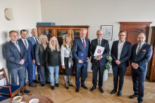 Dziesięć osób stojących w gabinecie. Pomieszczenie ma jasne ściany i drewnianą podłogę. W tle znajduje się regał z książkami i szafka. Na ścianie wisi obrazek. Osoby są elegancko ubrane – większość ma na sobie garnitury lub formalne stroje. Jedna osoba trzyma niebieską teczkę. Druga osoba trzyma białą teczkę z czerwonym herbem.