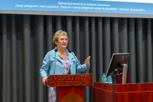 A woman standing behind a wooden lectern. She is dressed in a light blue jacket and a colorful blouse. To the left of the rostrum is a table on which a Dell laptop and several bottles of mineral water are placed. Dark curtains are visible in the background. At the top of the photo is a projection screen with the title of the event displayed.